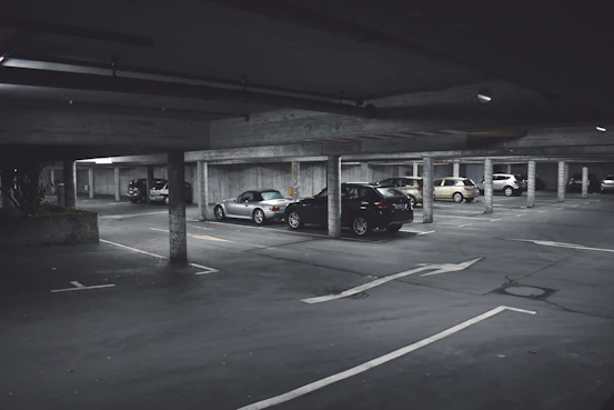 A dimly lit parking garage with several cars parked in marked spaces. Concrete pillars and overhead beams are visible, with directional arrows on the ground indicating traffic flow. The scene appears somewhat deserted with no visible people around.