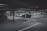 A dimly lit parking garage with several cars parked in marked spaces. Concrete pillars and overhead beams are visible, with directional arrows on the ground indicating traffic flow. The scene appears somewhat deserted with no visible people around.