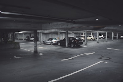 A dimly lit parking garage with several cars parked in marked spaces. Concrete pillars and overhead beams are visible, with directional arrows on the ground indicating traffic flow. The scene appears somewhat deserted with no visible people around.
