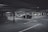 A dimly lit parking garage with several cars parked in marked spaces. Concrete pillars and overhead beams are visible, with directional arrows on the ground indicating traffic flow. The scene appears somewhat deserted with no visible people around.