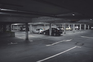 A dimly lit parking garage with several cars parked in marked spaces. Concrete pillars and overhead beams are visible, with directional arrows on the ground indicating traffic flow. The scene appears somewhat deserted with no visible people around.