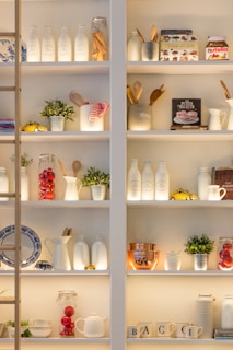 Shelves filled with various kitchen items, including jars, bottles, mugs, and small decorative plants. Notable objects include a book about baking, a Nutella jar, and small toy cars. The arrangement features a combination of culinary tools and decorative items like red glass spheres and plants, creating a homey and inviting atmosphere.