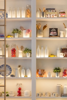 Shelves filled with various kitchen items, including jars, bottles, mugs, and small decorative plants. Notable objects include a book about baking, a Nutella jar, and small toy cars. The arrangement features a combination of culinary tools and decorative items like red glass spheres and plants, creating a homey and inviting atmosphere.