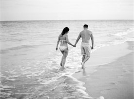 A couple is walking hand in hand along the shore, with gentle waves lapping at their feet. They are both wearing swimwear, and their footprints are visible in the wet sand. The ocean stretches into the distance under a light sky, conveying a sense of tranquility.