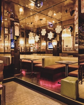 A cozy interior of a bar or restaurant with booths and tables, illuminated by warm lighting. Walls are adorned with framed artwork and photographs. Hanging lamps and decorative snowflakes add to the ambiance. A disco ball is visible on the ceiling, reflecting lights.