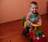 A young child is smiling while sitting on a colorful ride-on toy car in a room with wooden flooring and peach-colored walls. The child is wearing a striped long-sleeve shirt underneath a sleeveless zip-up top.