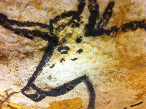 An ancient cave painting depicts the outline of a wild animal, possibly a prehistoric bison, with distinct horns and spots on its body. The artwork is on a textured rock surface with earthy tones.