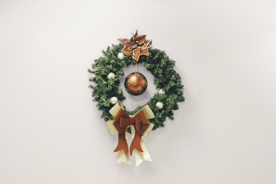 A festive wreath adorned with a golden poinsettia at the top, a large decorative bauble in the center, and smaller ornaments around the greenery. Additionally, a large golden and brown bow is attached at the bottom.
