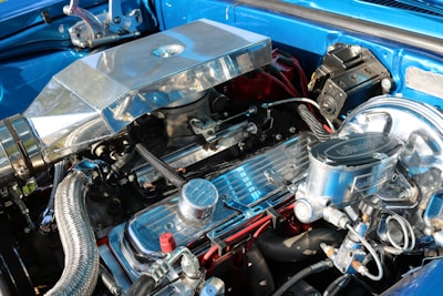 An intricately detailed car engine with polished metallic surfaces, including various tubes, hoses, and components. The engine is housed within a bright blue compartment and features shiny chrome finishes, suggesting high performance and customization.