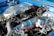 An intricately detailed car engine with polished metallic surfaces, including various tubes, hoses, and components. The engine is housed within a bright blue compartment and features shiny chrome finishes, suggesting high performance and customization.