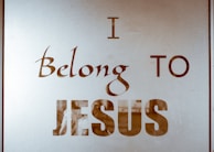 The image contains text on a reflective or translucent surface. The text is stylized and reads 'I Belong to Jesus' in bold and elegant typography. The background appears minimalistic, possibly due to the reflective nature of the surface.