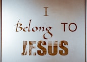 The image contains text on a reflective or translucent surface. The text is stylized and reads 'I Belong to Jesus' in bold and elegant typography. The background appears minimalistic, possibly due to the reflective nature of the surface.