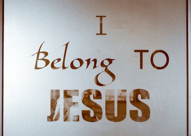 The image contains text on a reflective or translucent surface. The text is stylized and reads 'I Belong to Jesus' in bold and elegant typography. The background appears minimalistic, possibly due to the reflective nature of the surface.