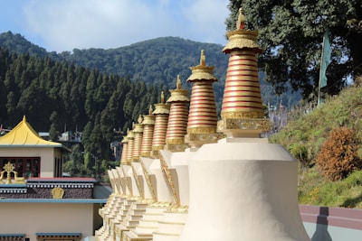 A row of intricately designed golden stupas with red accents, set against a backdrop of lush green hills and tall evergreen trees. The architecture features ornate details with a serene and peaceful monastery-like setting.