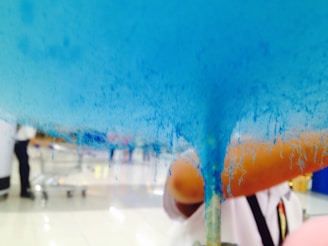 Blue cotton candy being made, with strands forming and a fuzzy texture. A person in the background, wearing a white shirt, is partially visible. The scene appears to be indoors, possibly in a shopping area or food court.