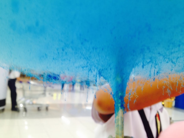 Blue cotton candy being made, with strands forming and a fuzzy texture. A person in the background, wearing a white shirt, is partially visible. The scene appears to be indoors, possibly in a shopping area or food court.