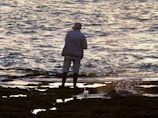 A person wearing a hat and jacket stands on a rocky shoreline while fishing, with the ocean waves gently splashing around them. The lighting suggests early morning or late afternoon, creating a peaceful and reflective atmosphere.