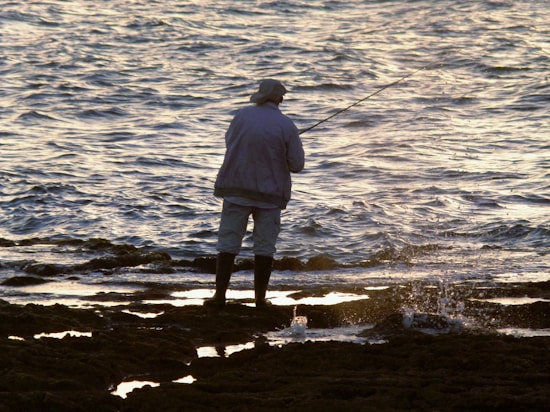 A person wearing a hat and jacket stands on a rocky shoreline while fishing, with the ocean waves gently splashing around them. The lighting suggests early morning or late afternoon, creating a peaceful and reflective atmosphere.