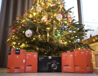 A festive Christmas tree adorned with colorful ornaments and glowing lights is prominently displayed. Below the tree, several boxed camera lenses and equipment from Sony are neatly arranged, suggesting a theme of photography gifts or presents.