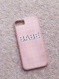 A pink phone case with a grid pattern lies on a textured carpet. The word 'BABE' is printed in bold, black letters across the center.