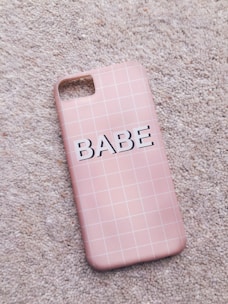A pink phone case with a grid pattern lies on a textured carpet. The word 'BABE' is printed in bold, black letters across the center.