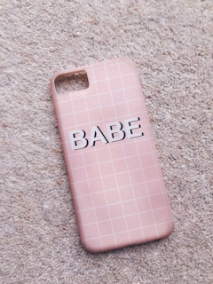 A pink phone case with a grid pattern lies on a textured carpet. The word 'BABE' is printed in bold, black letters across the center.