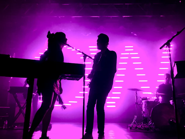 A colorful psychedelic backdrop with silhouettes of a band performing live.
