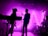 Silhouetted musicians perform on stage with a vibrant pink and purple backlight, creating an electric and energetic atmosphere. The figures of the band members and their instruments are visible against the illuminated backdrop.