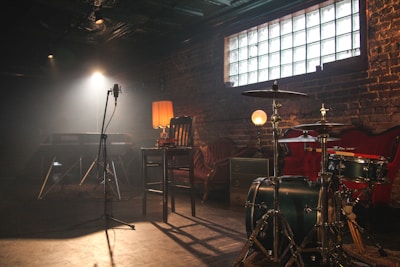 A dimly lit rustic music studio with various musical instruments and equipment. A drum set is positioned in the foreground, with a microphone stand centered in front of a keyboard. The background features exposed brick walls, an upholstered couch, a table lamp, and a soft glowing orb lamp, creating a warm and cozy ambiance.