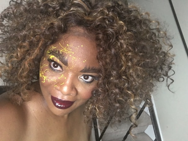 A person with curly hair is looking into the camera with bold makeup. Their face is decorated with yellow paint splatters, and their lips are painted a deep burgundy color.