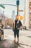 A man stands on a city street corner, dressed in a brown leather jacket with a shearling collar, jeans, and brown boots. Behind him, a busy urban scene unfolds with cars and pedestrians. Street signs indicating Central Park West and W 72 St are visible, hinting at a location in New York City. The atmosphere is clear with a hint of steam rising from a vent in the sidewalk, suggesting a chilly day.