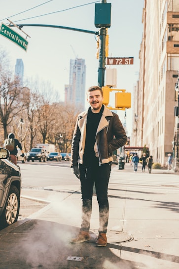 A man stands on a city street corner, dressed in a brown leather jacket with a shearling collar, jeans, and brown boots. Behind him, a busy urban scene unfolds with cars and pedestrians. Street signs indicating Central Park West and W 72 St are visible, hinting at a location in New York City. The atmosphere is clear with a hint of steam rising from a vent in the sidewalk, suggesting a chilly day.