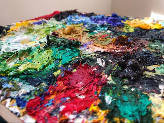 A close-up of a painter's palette with an assortment of multicolored oil paints. The paints are thickly applied and mixed together, creating various textures and shades. The dominant colors include vibrant greens, blues, reds, yellows, and blacks, all smeared and dolloped across the palette.
