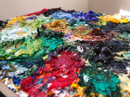 A close-up of a painter's palette with an assortment of multicolored oil paints. The paints are thickly applied and mixed together, creating various textures and shades. The dominant colors include vibrant greens, blues, reds, yellows, and blacks, all smeared and dolloped across the palette.