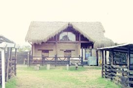 A rustic, thatched-roof building stands at the center, constructed mainly of wood and bricks. The surrounding area features wooden fences and grassy patches, giving the impression of a farm or rural setting. Some metal barrels and storage containers are visible near the structure.