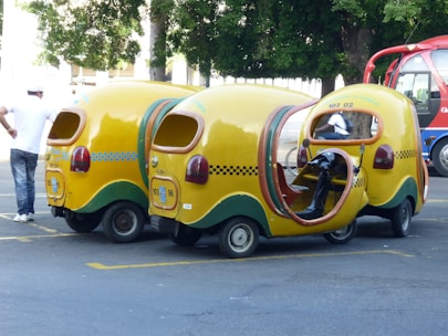 Three whimsical, yellow three-wheeled vehicles with vibrant cab-style designs and open sides are parked on the street. These vehicles have checkerboard patterns and green accents. A person in casual attire stands nearby, while another individual is seated inside one of the vehicles. A red and blue bus is visible in the background along with trees.