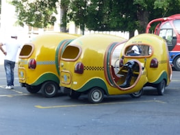 Three whimsical, yellow three-wheeled vehicles with vibrant cab-style designs and open sides are parked on the street. These vehicles have checkerboard patterns and green accents. A person in casual attire stands nearby, while another individual is seated inside one of the vehicles. A red and blue bus is visible in the background along with trees.
