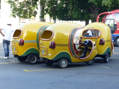 Three whimsical, yellow three-wheeled vehicles with vibrant cab-style designs and open sides are parked on the street. These vehicles have checkerboard patterns and green accents. A person in casual attire stands nearby, while another individual is seated inside one of the vehicles. A red and blue bus is visible in the background along with trees.