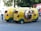 Three whimsical, yellow three-wheeled vehicles with vibrant cab-style designs and open sides are parked on the street. These vehicles have checkerboard patterns and green accents. A person in casual attire stands nearby, while another individual is seated inside one of the vehicles. A red and blue bus is visible in the background along with trees.