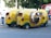 Three whimsical, yellow three-wheeled vehicles with vibrant cab-style designs and open sides are parked on the street. These vehicles have checkerboard patterns and green accents. A person in casual attire stands nearby, while another individual is seated inside one of the vehicles. A red and blue bus is visible in the background along with trees.
