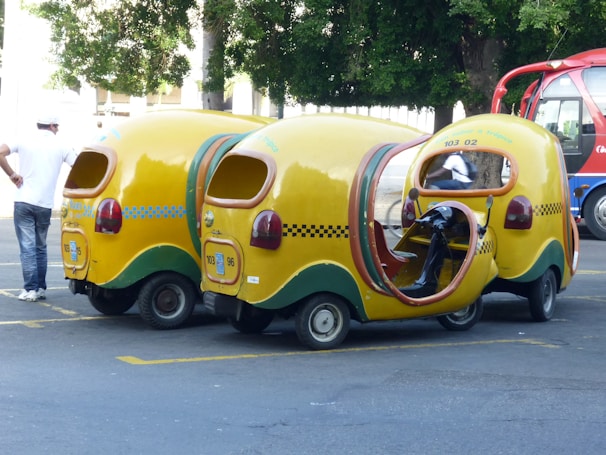 Three whimsical, yellow three-wheeled vehicles with vibrant cab-style designs and open sides are parked on the street. These vehicles have checkerboard patterns and green accents. A person in casual attire stands nearby, while another individual is seated inside one of the vehicles. A red and blue bus is visible in the background along with trees.