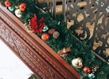 A decorative garland adorned with various ornaments including red, green, and gold baubles, pinecones, and a large red flower. It's placed on a carved wooden banister, adding a festive touch to the setting. The background features intricately designed black metal railings.