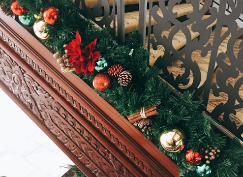 A decorative garland adorned with various ornaments including red, green, and gold baubles, pinecones, and a large red flower. It's placed on a carved wooden banister, adding a festive touch to the setting. The background features intricately designed black metal railings.