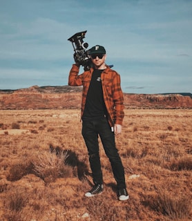 A person stands in a dry, open landscape with a mountainous backdrop, holding a professional camera on their shoulder. The individual is wearing a black cap, sunglasses, a brown plaid shirt, black pants, and black sneakers.