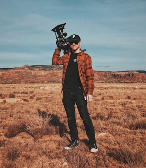 A person stands in a dry, open landscape with a mountainous backdrop, holding a professional camera on their shoulder. The individual is wearing a black cap, sunglasses, a brown plaid shirt, black pants, and black sneakers.