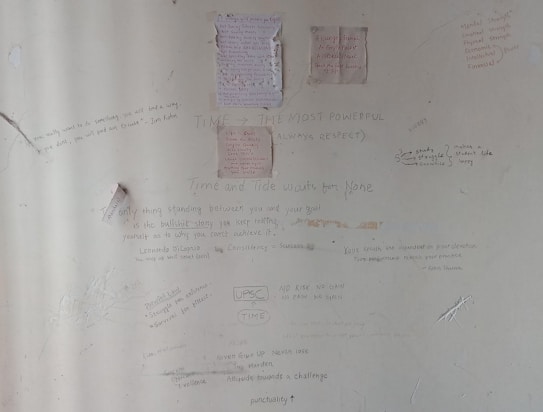 A wall covered in various handwritten motivational quotes and notes. Some quotes are attributed to notable personalities and focus on themes like persistence, success, and self-belief. There are drawings and scribbles, including diagrams and small notes, emphasizing hard work and dedication.
