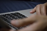 Fingers are positioned over the keyboard of a laptop, suggesting typing or interaction with the device. The keyboard displays symbols not commonly used in Western languages.