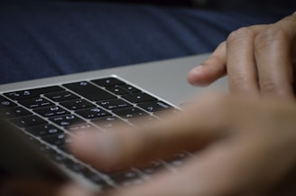 Fingers are positioned over the keyboard of a laptop, suggesting typing or interaction with the device. The keyboard displays symbols not commonly used in Western languages.