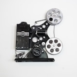A vintage film projector with two large reels and various mechanical components. The device is marked with the brand name 'Keystone' and features a classic, industrial design with metallic and black parts.