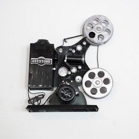A vintage film projector with two large reels and various mechanical components. The device is marked with the brand name 'Keystone' and features a classic, industrial design with metallic and black parts.
